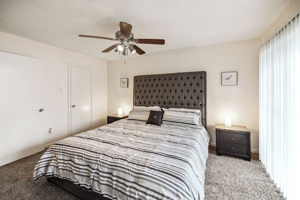 A bedroom with a tufted headboard, a striped bedspread, two bedside tables with lamps, a ceiling fan, and a door.