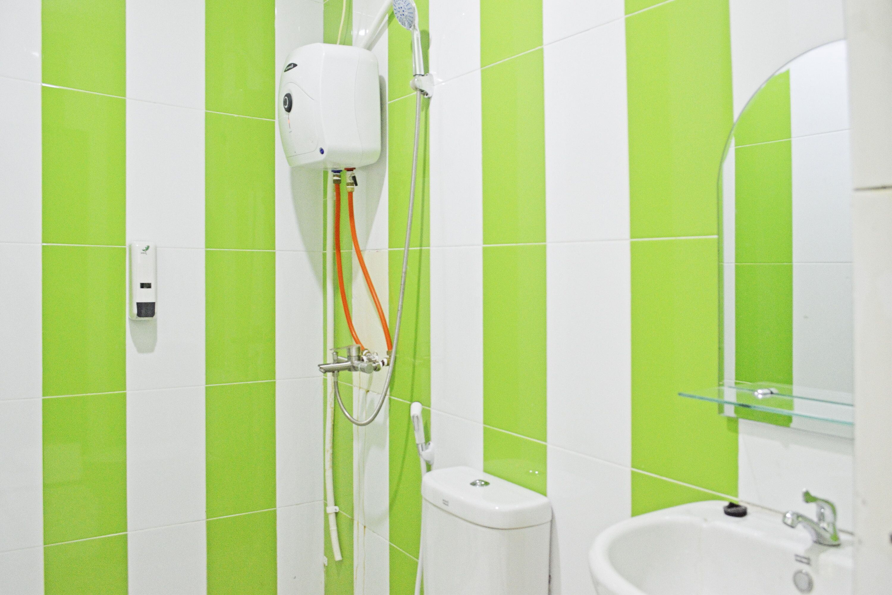 Un baño con azulejos blancos y verdes, un lavamanos blanco, un cabezal de ducha de mano y un estante de vidrio.