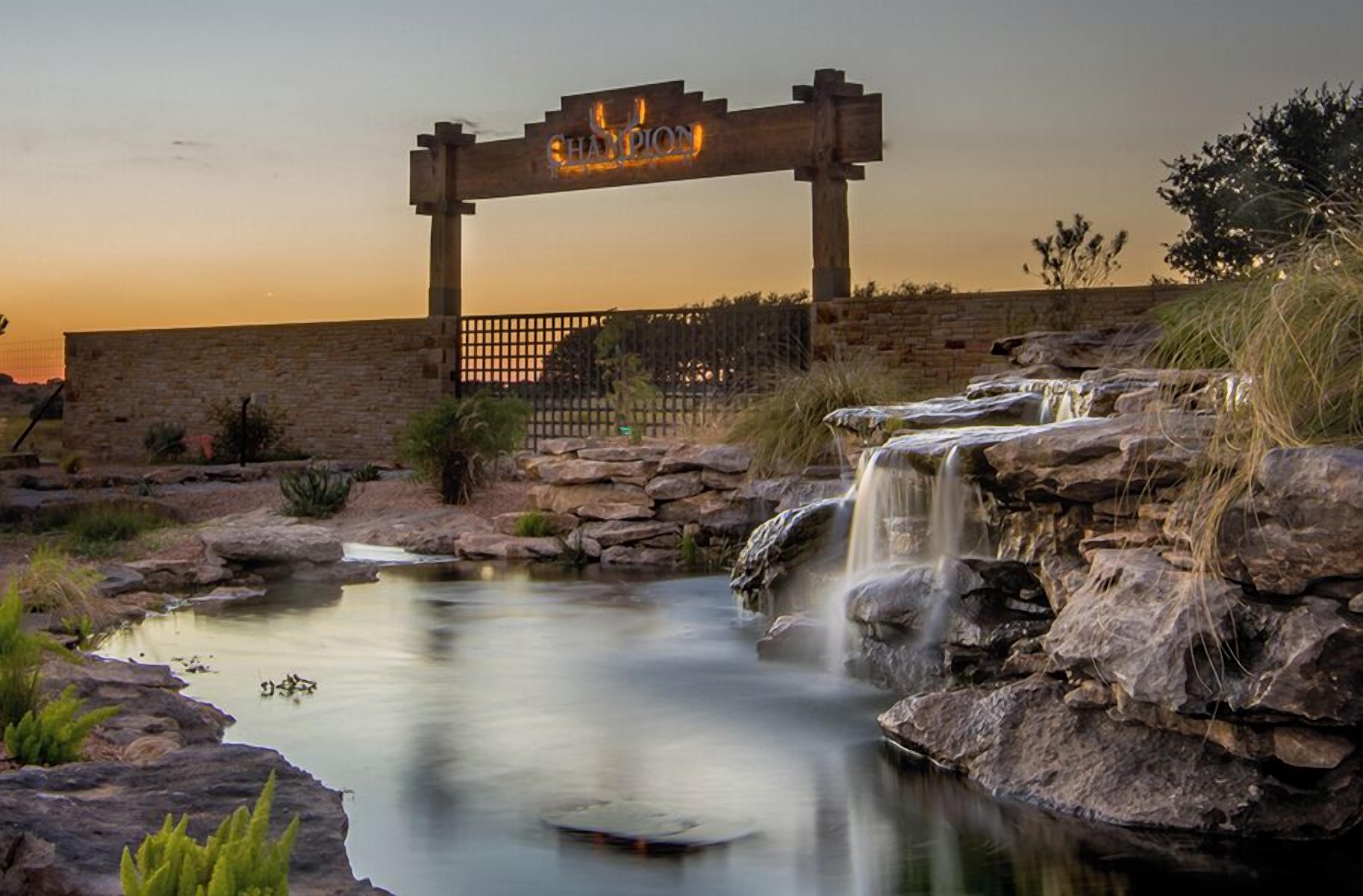 A wooden archway with the word "CHAMPION" illuminated, a small waterfall, and a calm pond with rocks and vegetation.