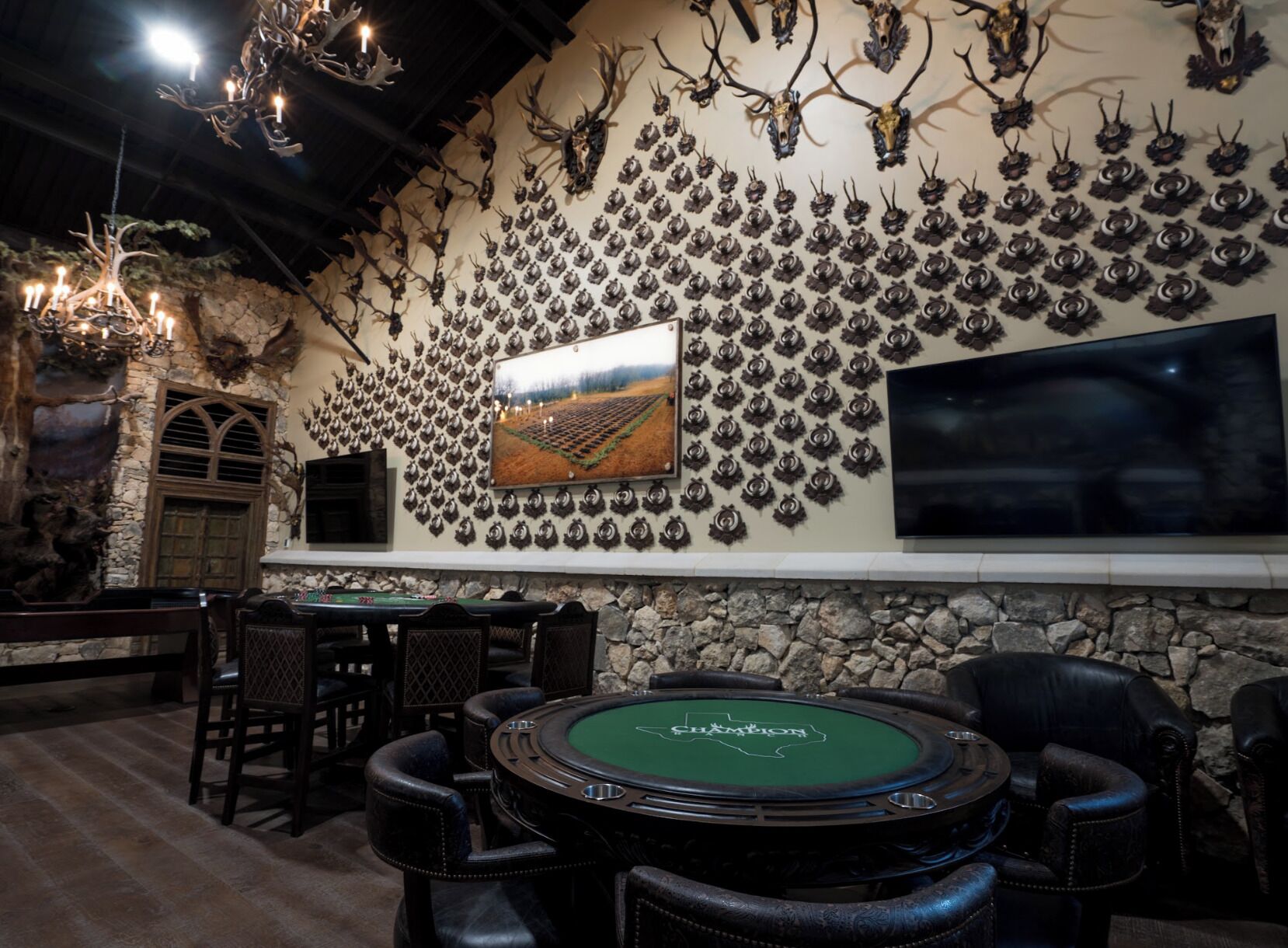 A casino with a green felt poker table, multiple chairs, a wall decorated with antlers and a painting, and a large TV screen.