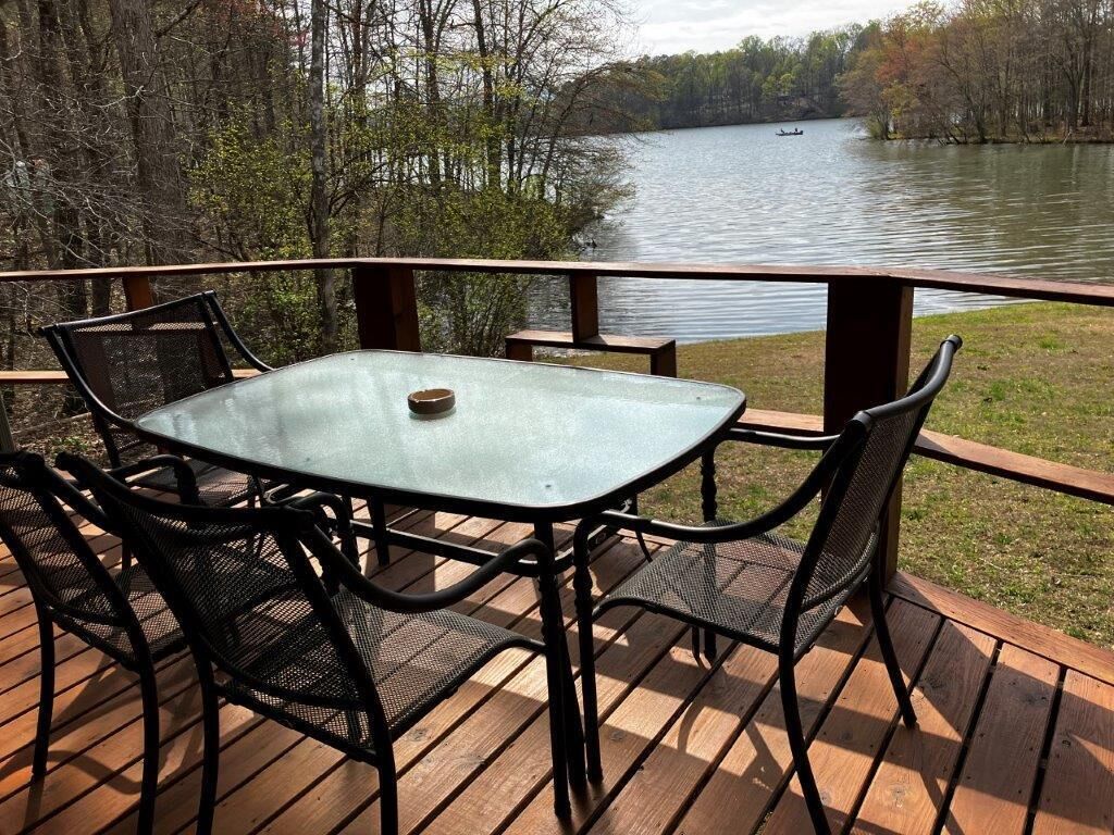 A deck with a glass table and chairs, overlooking a lake with a boat in the distance.