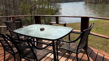 A deck with a glass table and chairs, overlooking a lake with a boat in the distance.