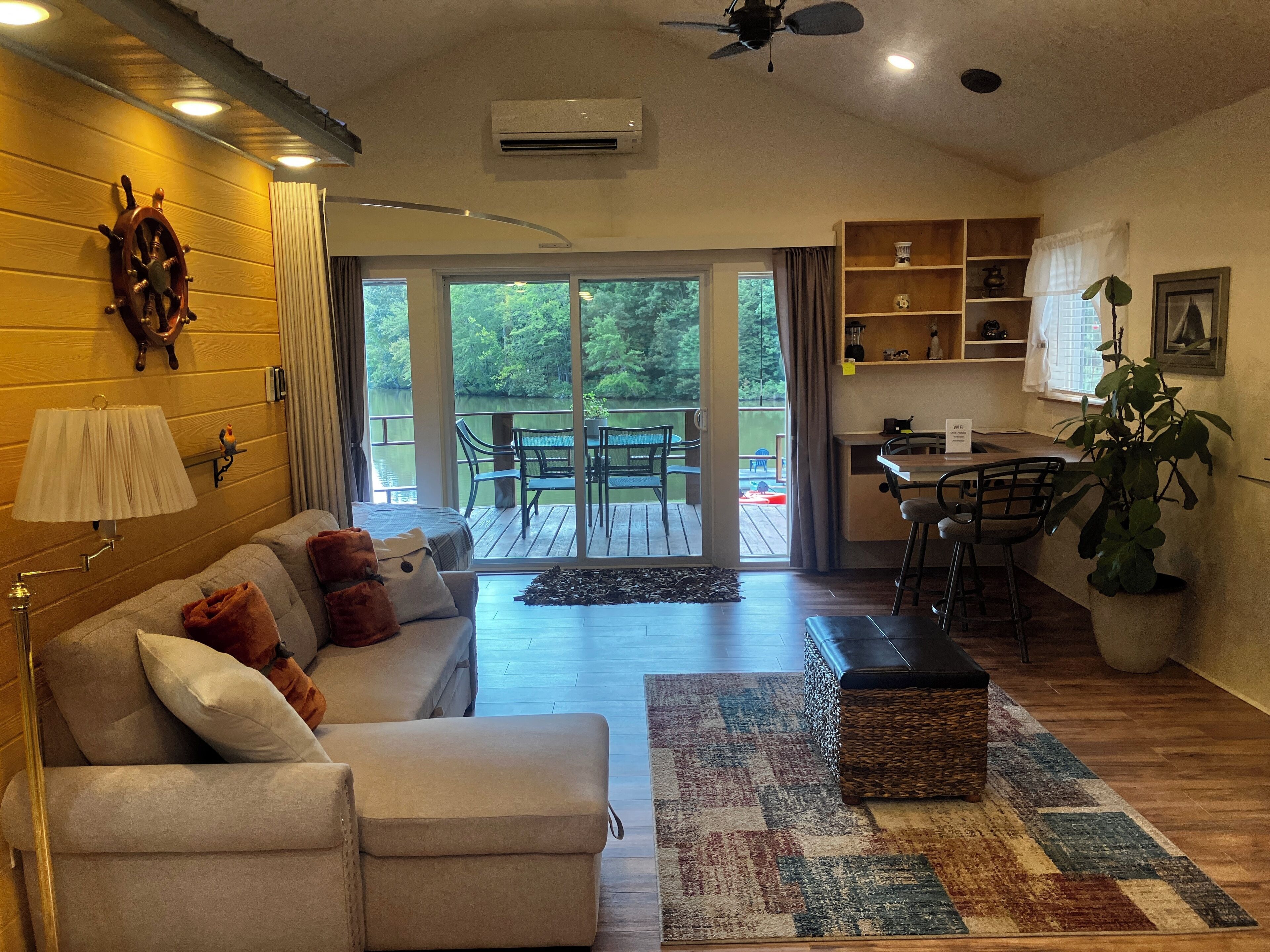 A cozy living room with a sofa, a coffee table, and a rug. There is a dining area with chairs and a table. A large window offers a view of a deck and greenery.
