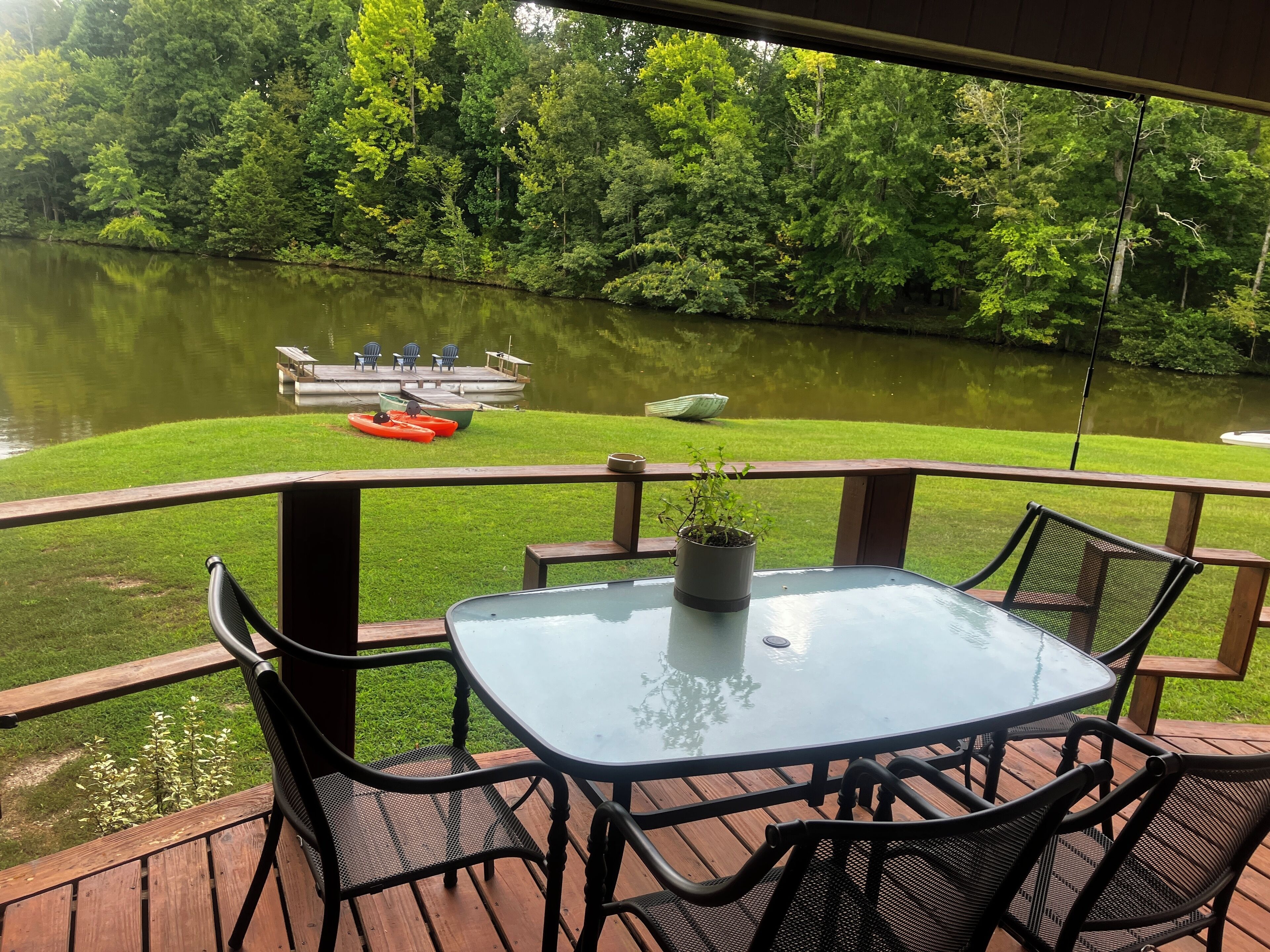 Een terras met een glazen tafel en stoelen, uitkijkend op een meer met boten en een steiger.