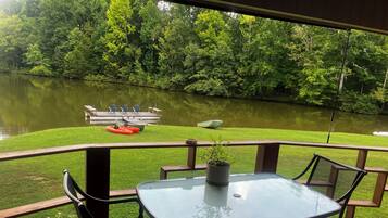 Een terras met een glazen tafel en stoelen, uitkijkend op een meer met boten en een steiger.