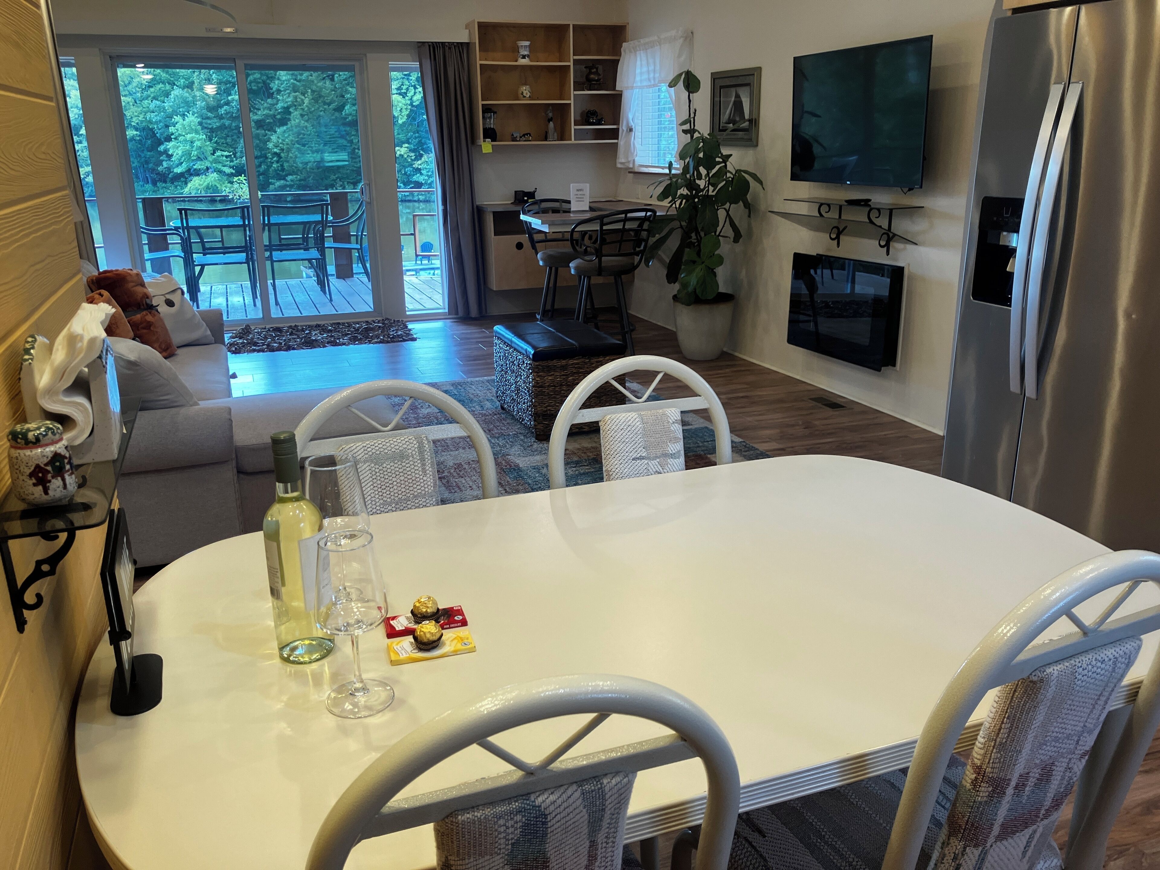 A dining area with a white table and chairs, a bottle of wine, and a plate of food. There is a TV mounted on the wall and a refrigerator to the right.