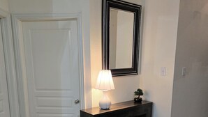 Een hal met een witte deur, een donkere console en een lamp, met daarboven een spiegel.