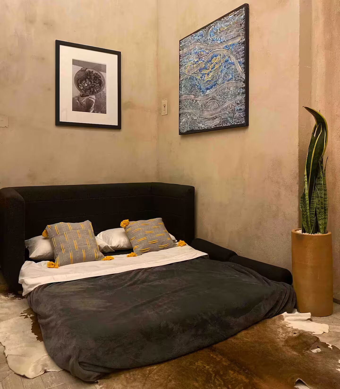 A bedroom with a black headboard, a bed with a grey and yellow patterned blanket, a framed picture on the wall, a large abstract painting, and a tall potted plant.