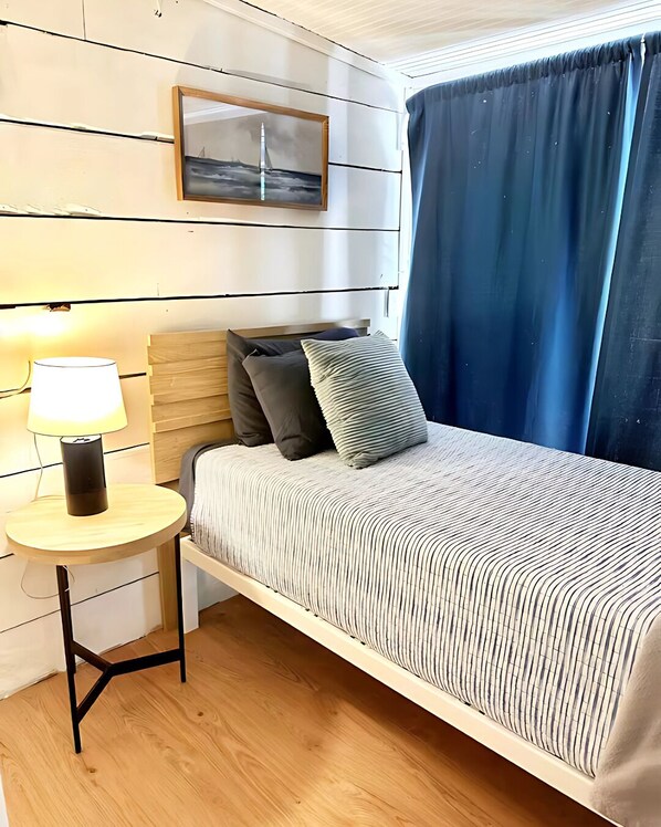 A bedroom with a bed, a bedside table with a lamp, a wall-mounted picture, and a window with curtains.