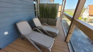 Ein Balkon mit zwei Liegestühlen, einer Holzterrasse, einer blauen Wand und Blick auf eine Wohngegend.