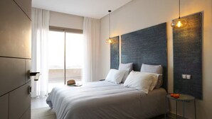 Ein modernes Hotelzimmer mit einem großen Bett, einem Nachttisch mit Lampe und Blick ins Freie durch ein großes Fenster.