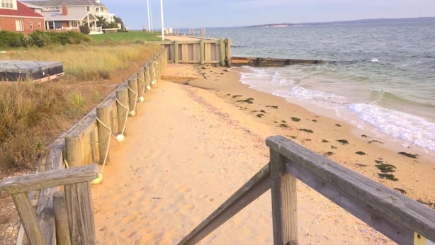 Een houten looppad dat naar een zandstrand leidt, met golven, een grasveld en een huis op de achtergrond.