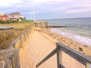 A wooden boardwalk leading to a sandy beach with waves, a grassy area, and a house in the background. - North Fork Summer Escape - 5BR Waterfront Home with Private Beach Access (Southhold)