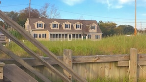 Een houten trap leidt naar een huis met een ruime veranda, omringd door hoog gras.