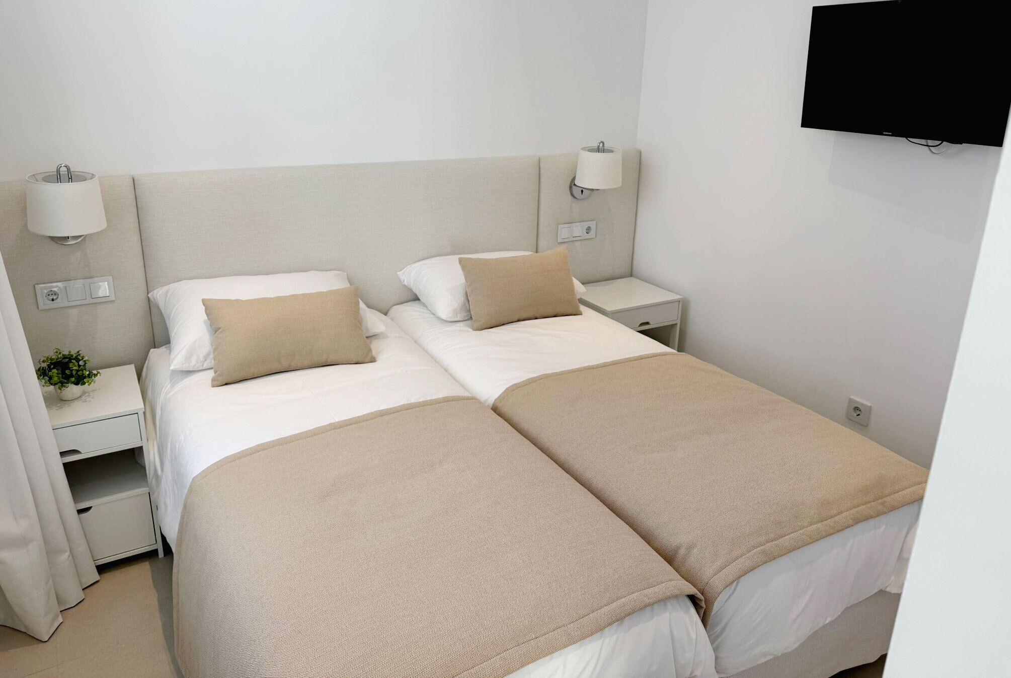 A hotel room with two beds, each with a beige blanket and pillows, a bedside table with a lamp, and a TV mounted on the wall.