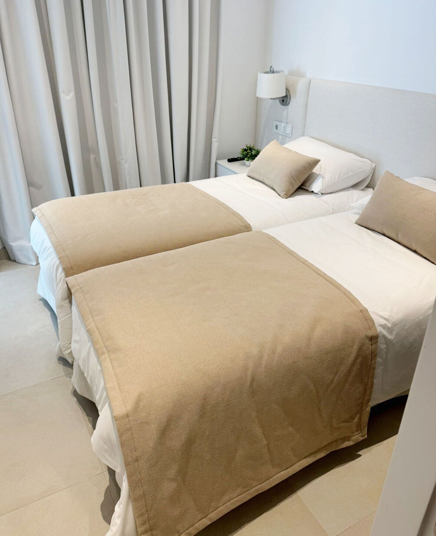 A hotel room with two beds, each with a beige bedspread and pillows. There is a nightstand with a lamp and a small plant.