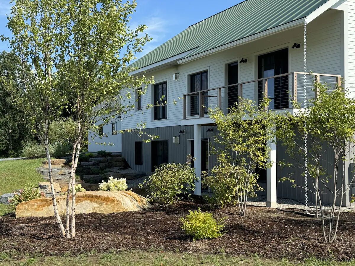 A two-story house with a green roof, a stone staircase leading to the entrance, and a well-maintained garden with young trees and shrubs.