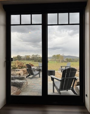 Vista a través de una puerta corrediza a un patio al aire libre con dos sillas Adirondack y una mesa, con vistas a un paisaje cubierto de césped y árboles bajo un cielo nublado.