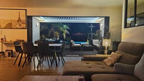 A modern living room with a sofa, dining table, and chairs. There is a view of a pool and outdoor area at night. - AIR-CONDITIONED VILLA WITH POOL IN PORTICCIO (Grosseto-Prugna)