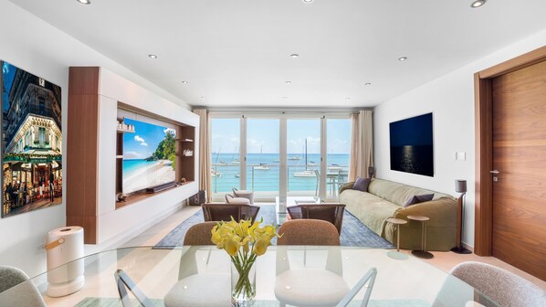 A modern living room with a glass dining table, comfortable seating, a large TV displaying a beach scene, and a view of the sea through large windows. - Luxurious 2BR/2.5BA Unit on Simpson Bay Beach (Simpson Bay)