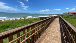 Ein hölzerner Steg führt zu einem Strand mit Sonnenschirmen und Menschen.