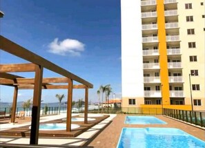 Et hotel med et poolområde, en træpergola og en høj bygning med altaner.