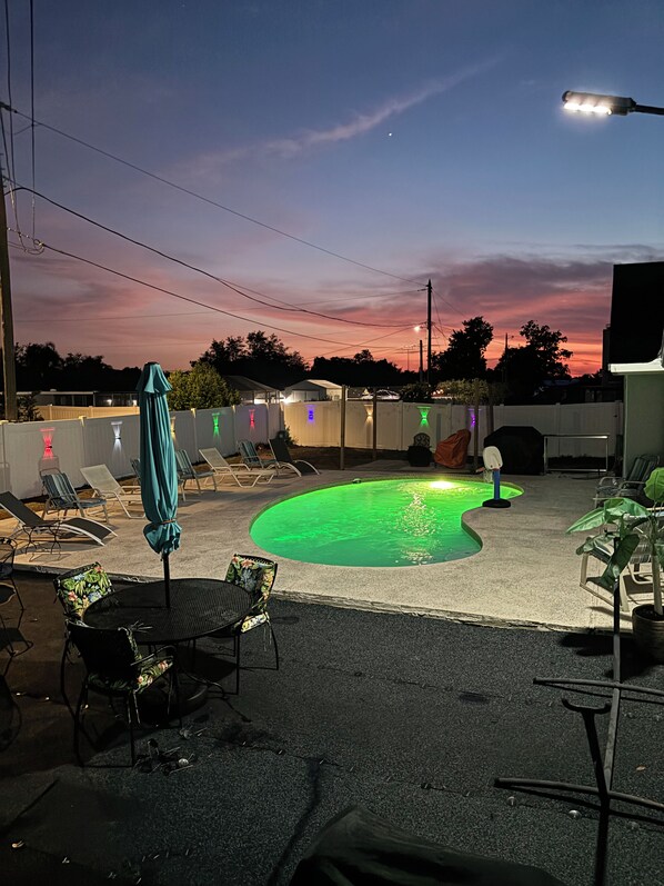 A swimming pool area with lounge chairs and umbrellas, surrounded by a fence and a building in the background. - Spacious 4-bedroom house in serene Deltona with AC, WiFi (Deltona)