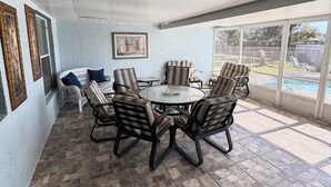 A covered patio with a stone-tiled floor, a round glass table surrounded by striped chairs, and a view of a swimming pool through large sliding glass doors. - Spacious 4-bedroom house in serene Deltona with AC, WiFi (Deltona)