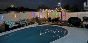 A backyard with a swimming pool, outdoor seating, and string lights.