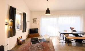 A modern living room with a TV, sofa, dining table, and a large window with curtains.