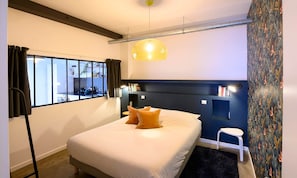 A modern hotel room with a bed, a small side table, a window with curtains, a ceiling light, and a patterned wall.