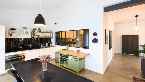 Una cocina moderna con una lámpara colgante negra, una isla verde y una mesa de comedor con un jarrón de flores.