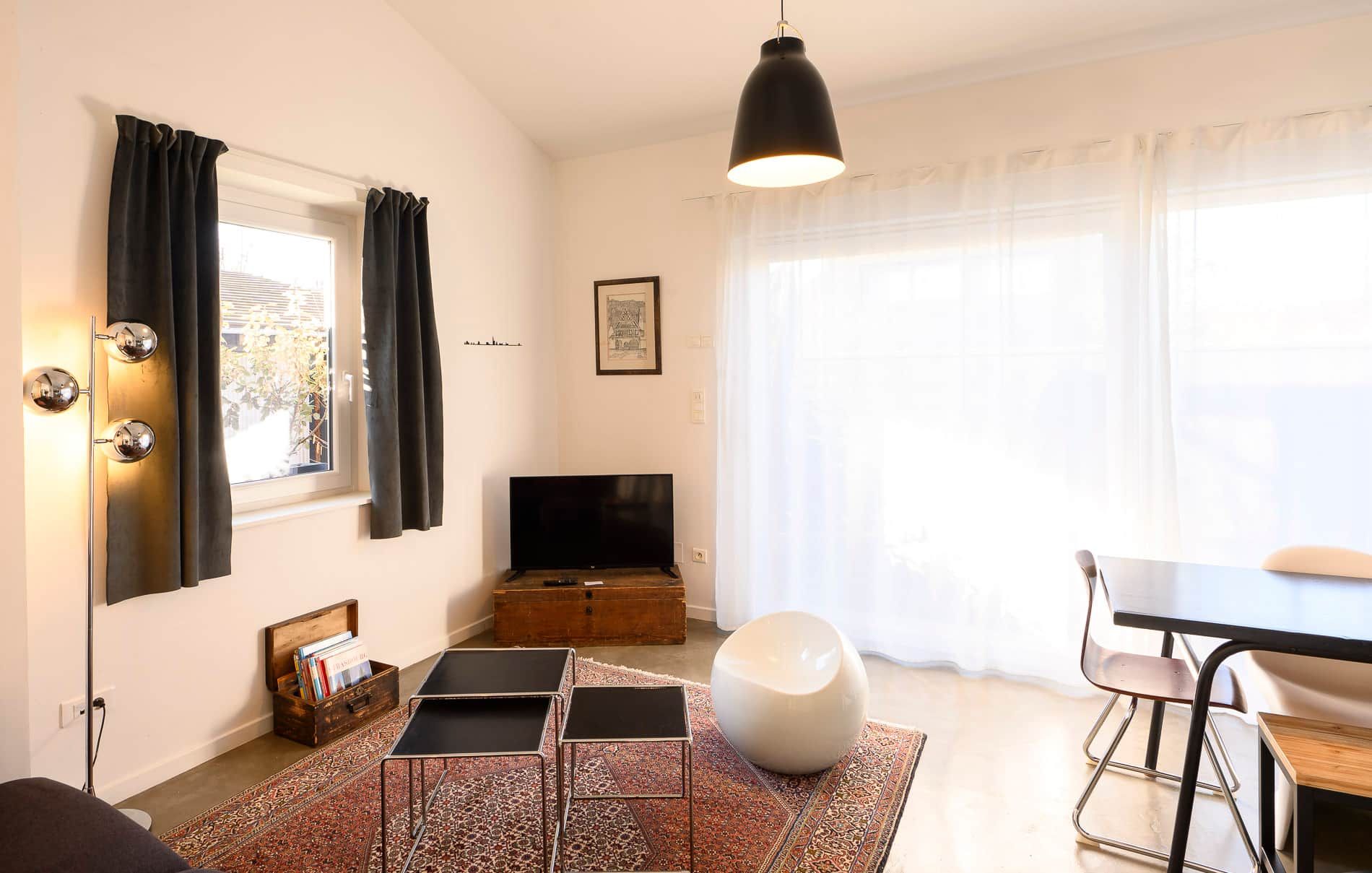 A modern living room with a flat-screen TV, a dining area with a table and chairs, a large window with sheer curtains, and a patterned rug.