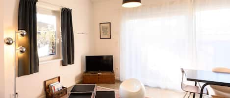 Una sala de estar moderna con un televisor de pantalla plana, un comedor con mesa y sillas, un ventanal con cortinas de gasa y una alfombra estampada.