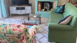 A living room with a green sofa, a floral ottoman, a flat-screen TV on a white cabinet, a fireplace with decorative items, and a patterned rug.