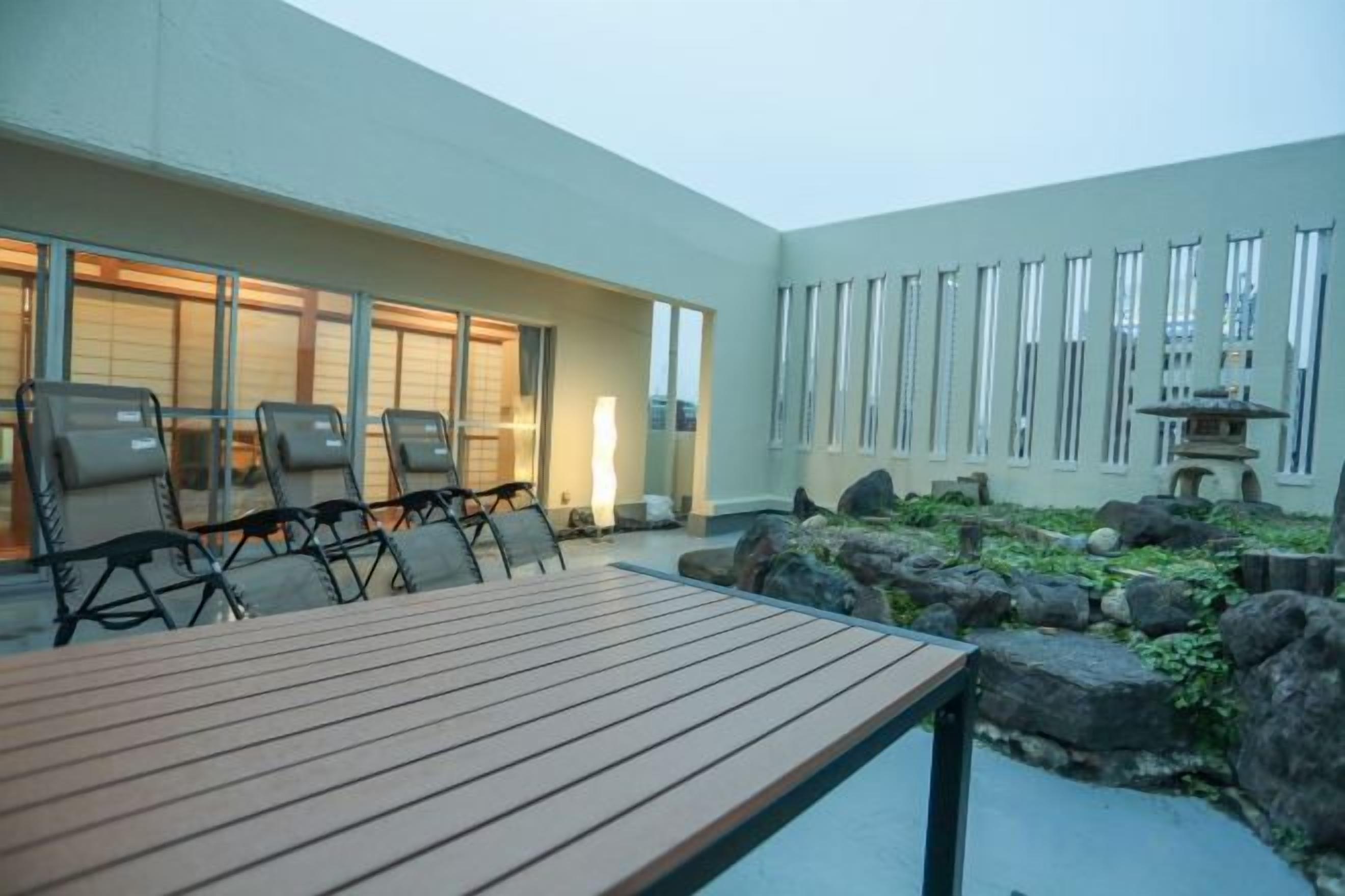 An outdoor seating area with a wooden table and chairs, a small rock garden, and a building with large windows.