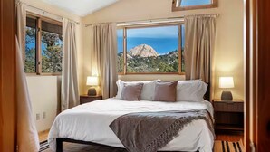 Un dormitorio con una cama grande, dos mesitas de noche con lámparas, vista a la montaña a través de una ventana y cortinas.