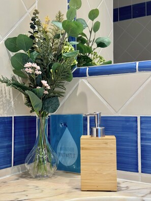 Un baño con un revestimiento de azulejos azul y blanco, un florero transparente con flores y un dispensador de jabón sobre un bloque de madera.