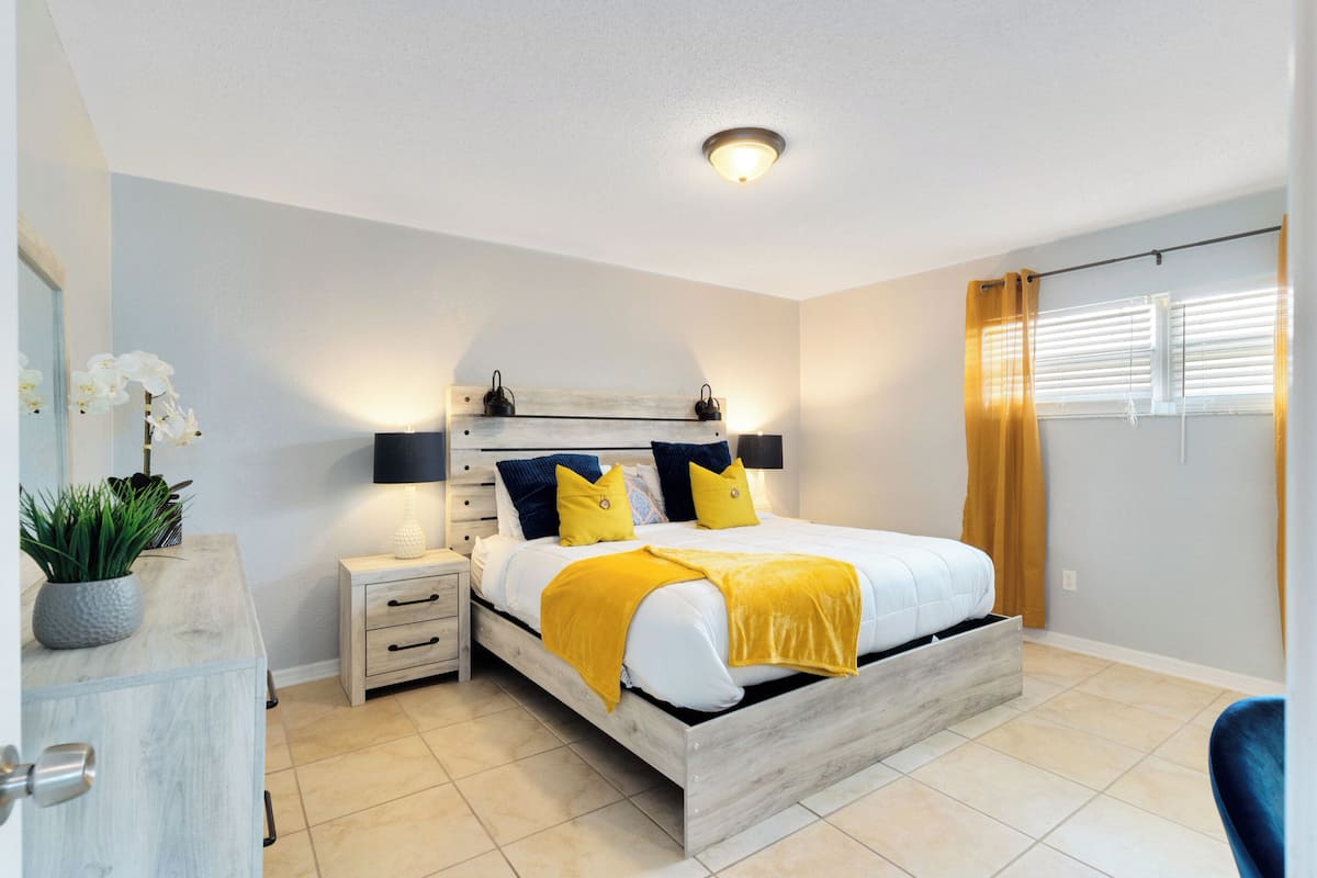 Un dormitorio con cama, mesitas de noche, cómoda, planta en maceta y ventana con cortinas.