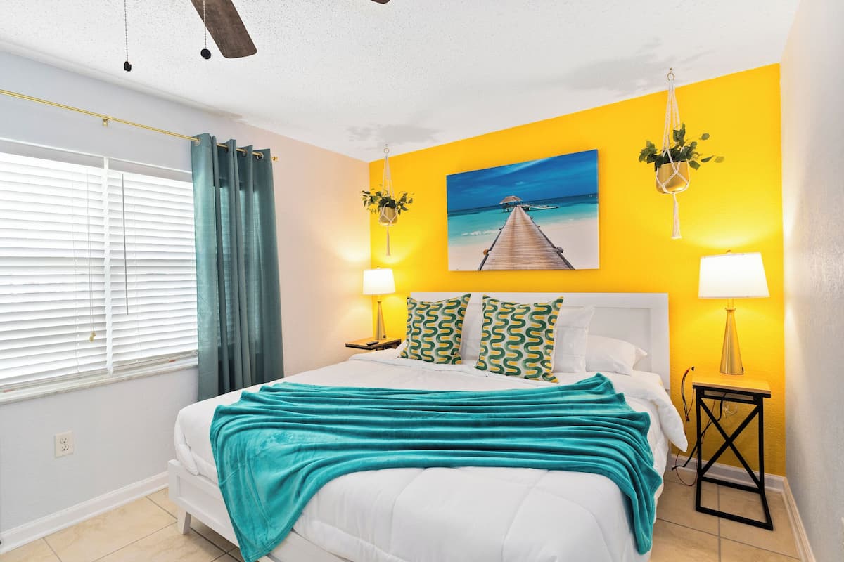Un dormitorio con una cama, un cuadro de un muelle, dos lámparas de noche y una ventana con cortinas.