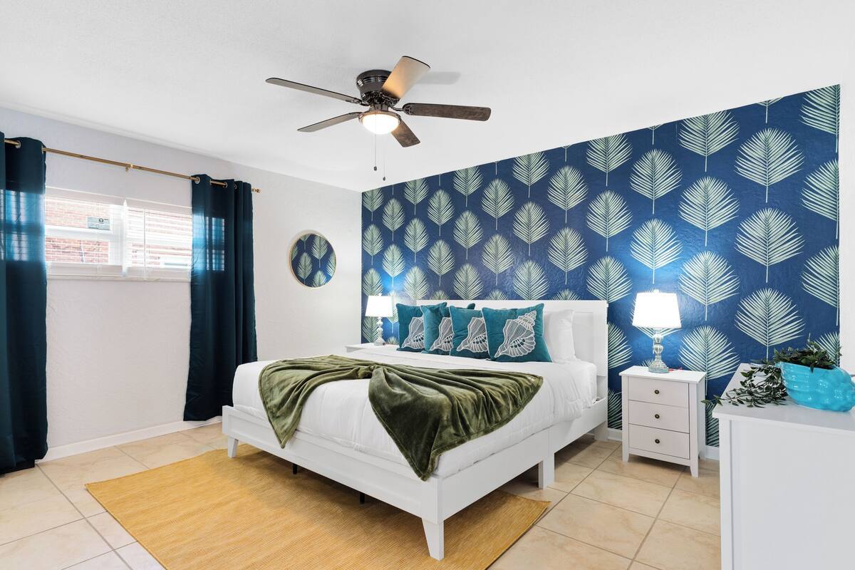 Un dormitorio con cama, mesita de noche, lámpara, ventilador de techo y ventana con cortinas.