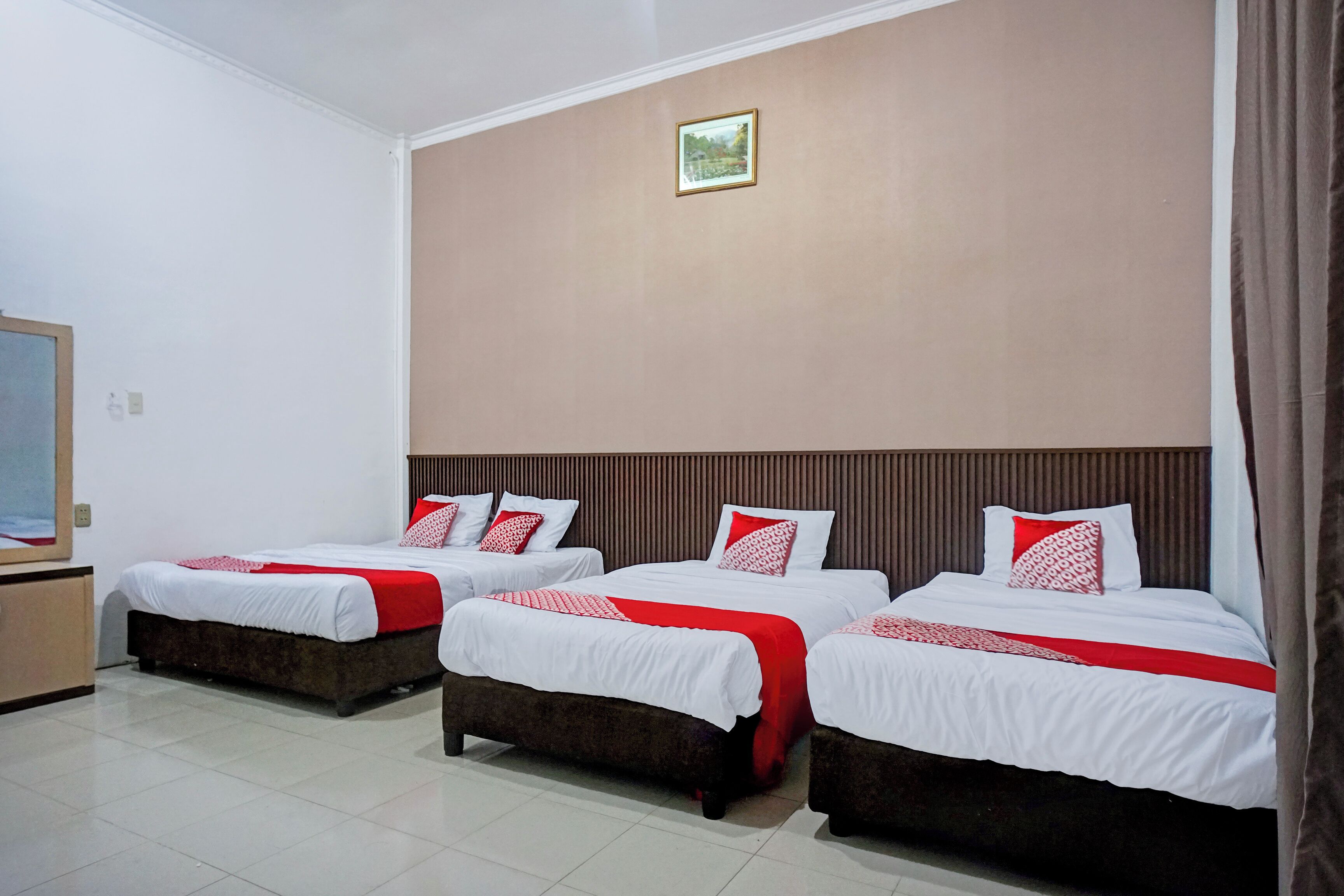 A hotel room with three beds, each with white bedding and red accents, a wooden headboard, and a small framed picture on the wall.