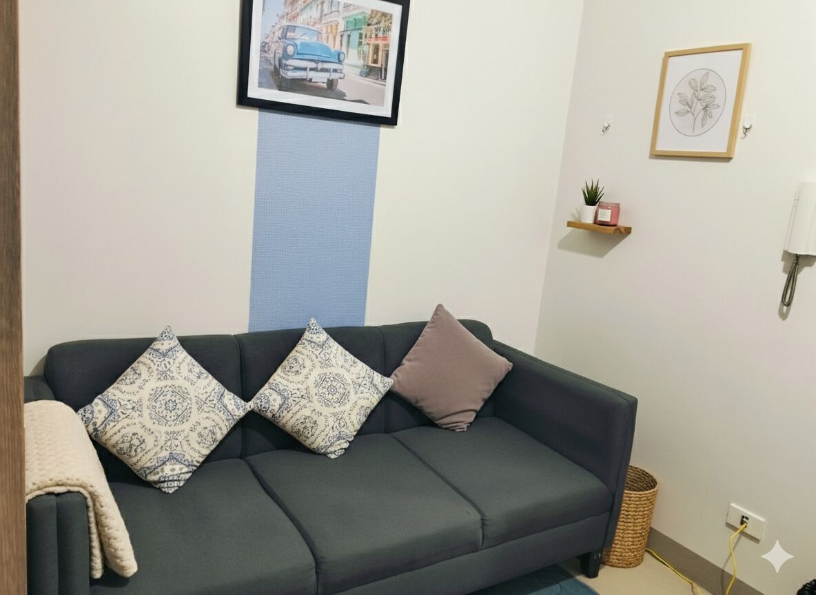Un salón con un sofá verde oscuro, cojines estampados, un cuadro enmarcado en la pared, un estante pequeño con una planta y una lámpara.