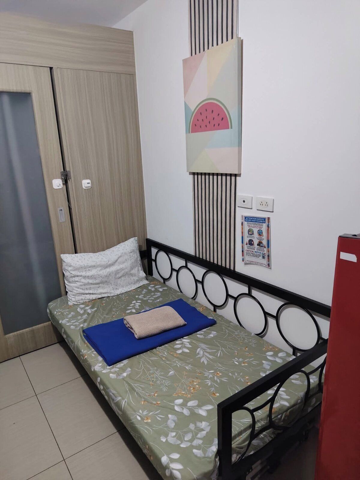 A single bed with a floral bedspread, a blue pillow, and a folded blanket. There is a wall-mounted artwork of a watermelon.