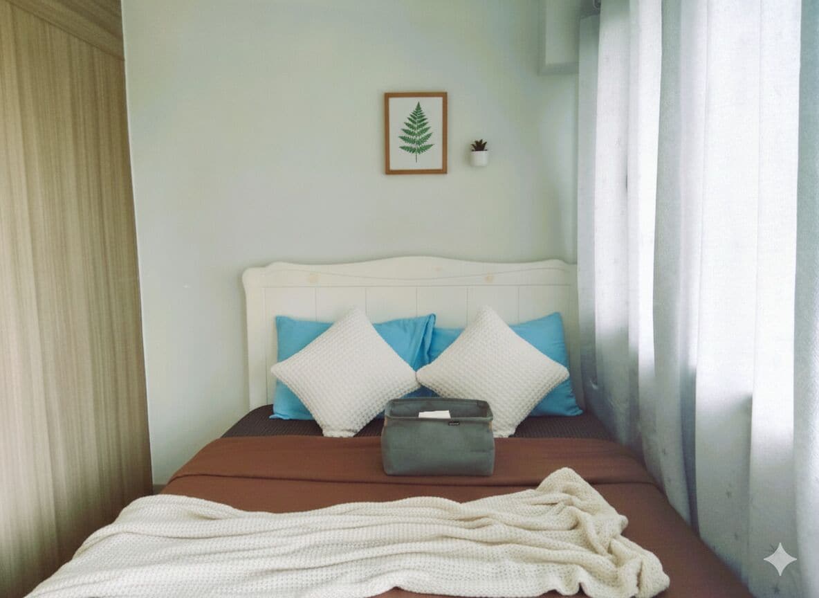 Un lit bien fait, avec un matelas marron, une tête de lit blanche et deux oreillers bleus. On aperçoit une table de chevet grise, sur laquelle se trouvent une petite plante et un décor en forme de feuille encadrée.