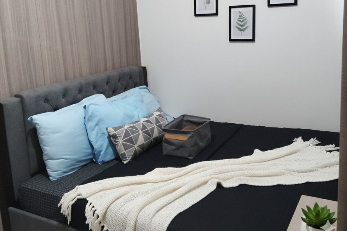Un dormitorio moderno con un cabecero gris capitonado, una colcha negra y una manta blanca. Hay cuatro láminas botánicas enmarcadas en la pared.