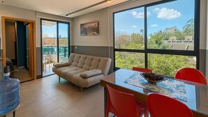 A modern living room with a sofa, dining table, and chairs, large windows with a city view, and a water dispenser. - Edifício Villa Oceânica  (Ipojuca)