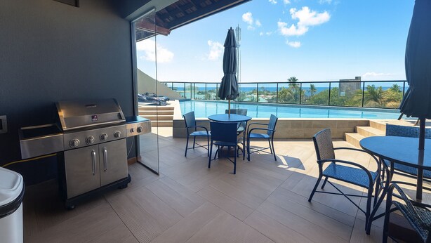 A balcony with a grill, chairs, and a view of the pool and ocean. - Edifício Villa Oceânica  (Ipojuca)