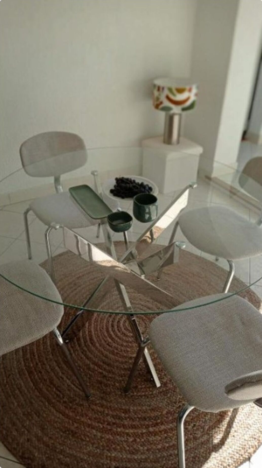 A round glass table with four chairs, a woven rug, and a white cabinet with a decorative item in the background.