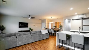 A modern living room with a grey sofa, wooden flooring, a dining area with a table and chairs, and a kitchen with a white countertop and bar stools. - 5 Bedroom House in Beautiful SW Bakersfield - HWY 99 Access (Bakersfield)